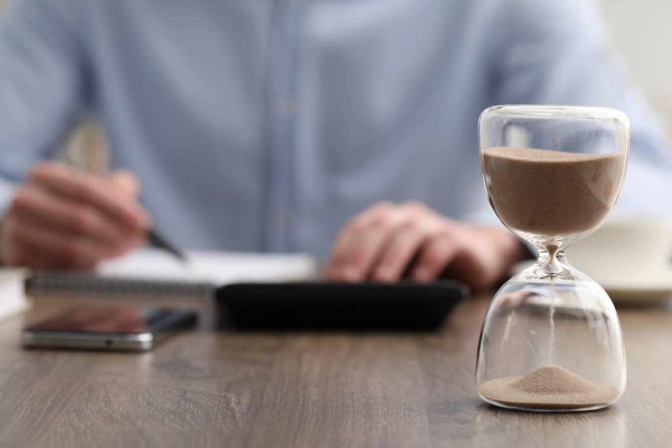 hourglass-with-flowing-sand-desk-man-taking-notes-while-using-calculator-indoors-selective-focus.jpg